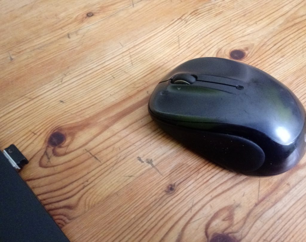 Une souris sans fil posée sur une table en bois. Sur la gauche de la photo, le bord d'un ordinateur, et le dungle usb associé à la souris.