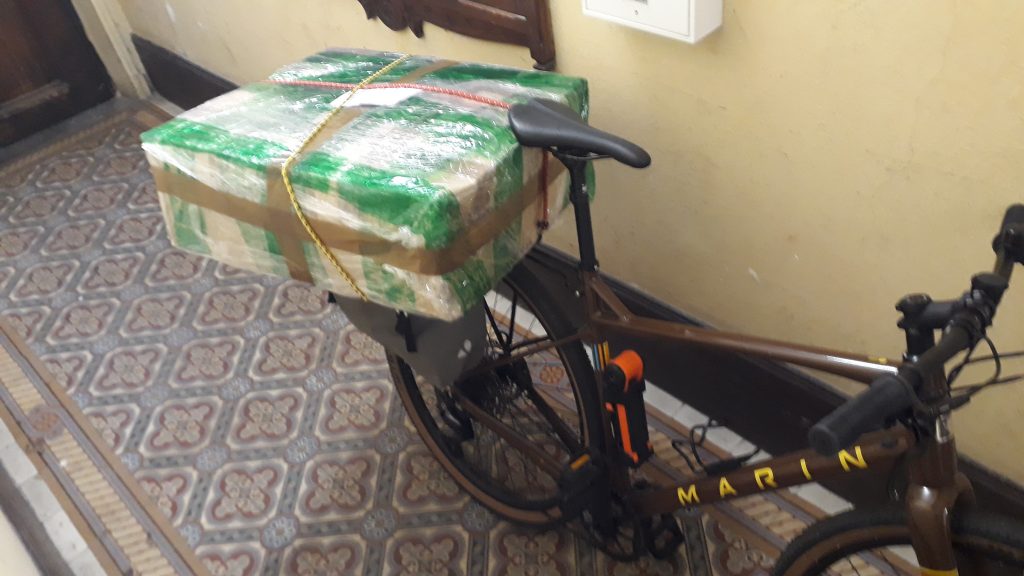 Un assez gros carton tenu par des tendeurs sur le bagage arrière du vélo.