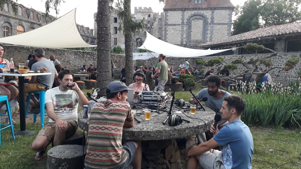 5 personnes en train de discuter au micro, autour d'une table en pierre. Derrière eux, des gens sont installés sur des tables. Au loin, le château.